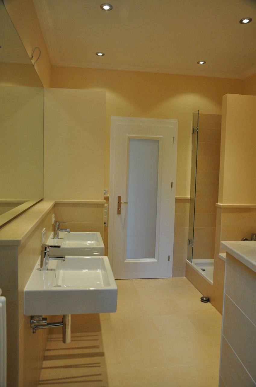 Badezimmer mit zwei Waschbecken, cremefarbenen Wänden über hellem Steinsockel und Duschkabine neben der Tür