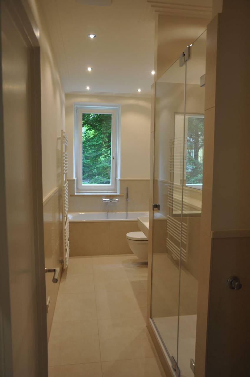 Schmales Badezimmer mit eingefasster Badewanne unter dem Fenster, Wand-WC und Duschkabine hinter Glas