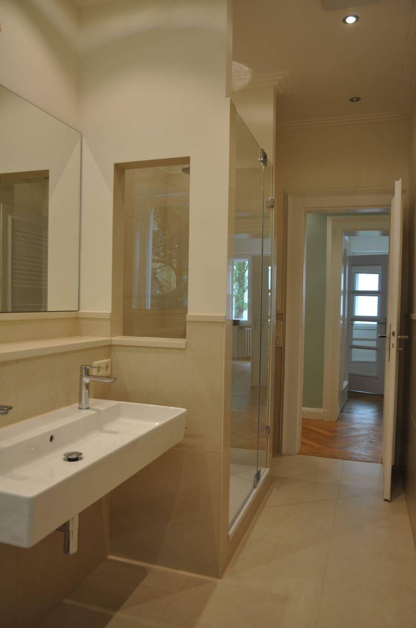 Badezimmer mit langem Waschbecken, wandhohem Spiegel, gefliester Ablagenische und Dusche hinter Glas