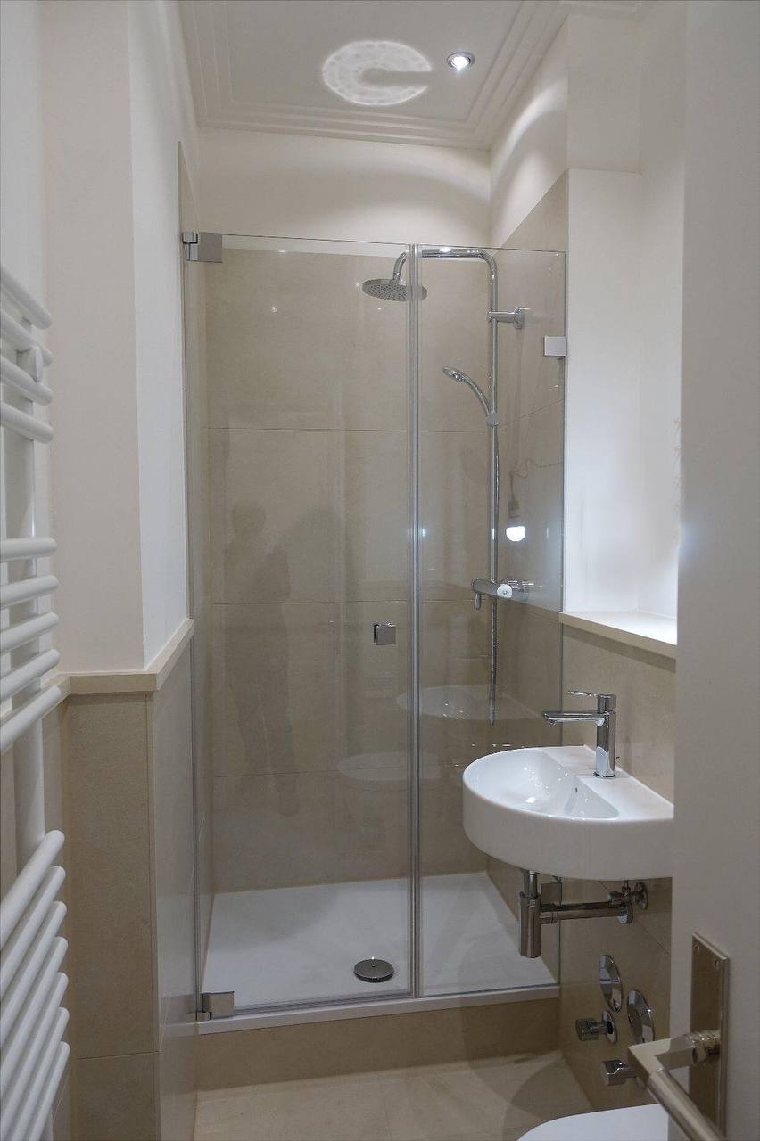 Dusche mit Glastür und Regenbrause, Wandflächen in hellem Stein, daneben ein Eckwaschbecken